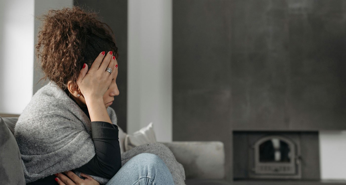 Woman wrapped in a blanket sits on couch holding her head