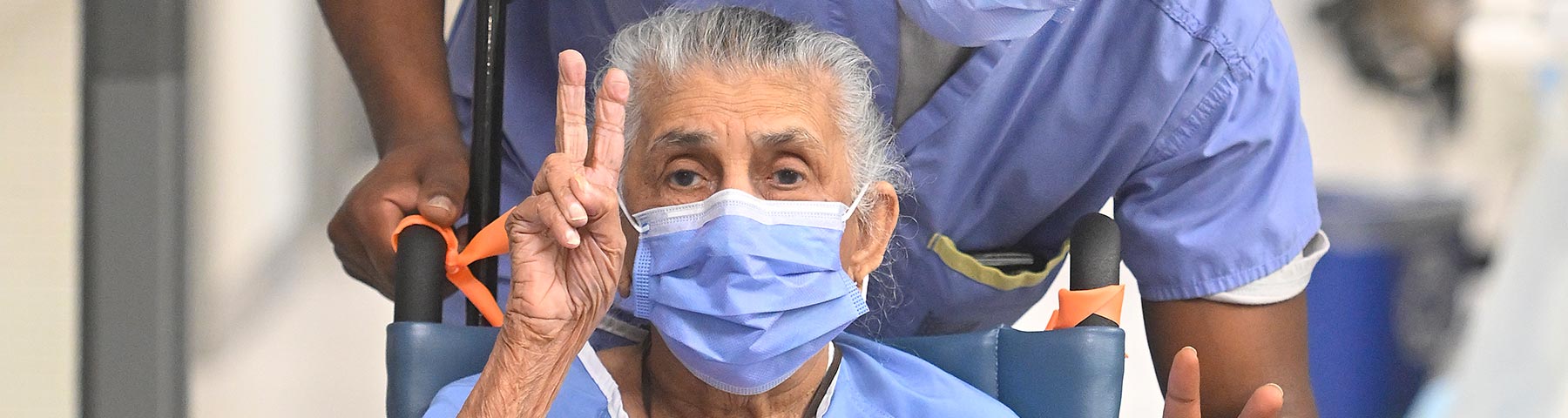 Older female patient in a wheelchair holding up a peace sign while being pushed down the hallway by a clinician